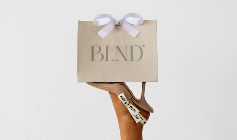 Beige shopping bag with 'BLND' branding held by a foot wearing a high-heeled shoe.