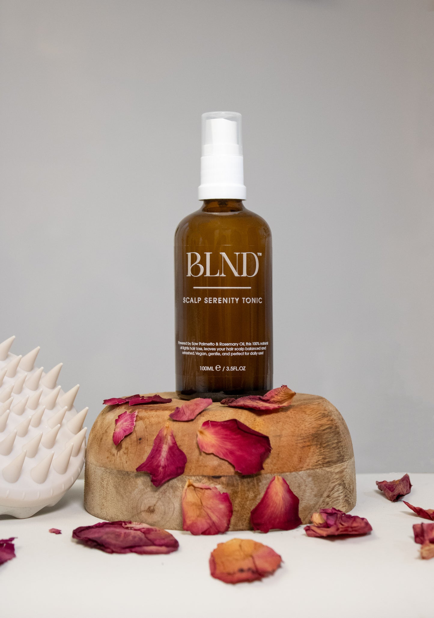 Bottle of BLND Scalp Serenity Tonic on a wooden block with rose petals, against a gray background.